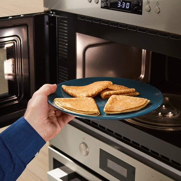 A person holding a blue plate with golden pancakes near a microwave oven.