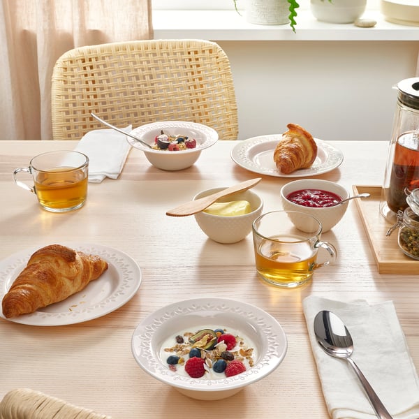 Wooden table set with glassware, yoghurt, berries, granola, croissants, and tea. Wicker chair in background.