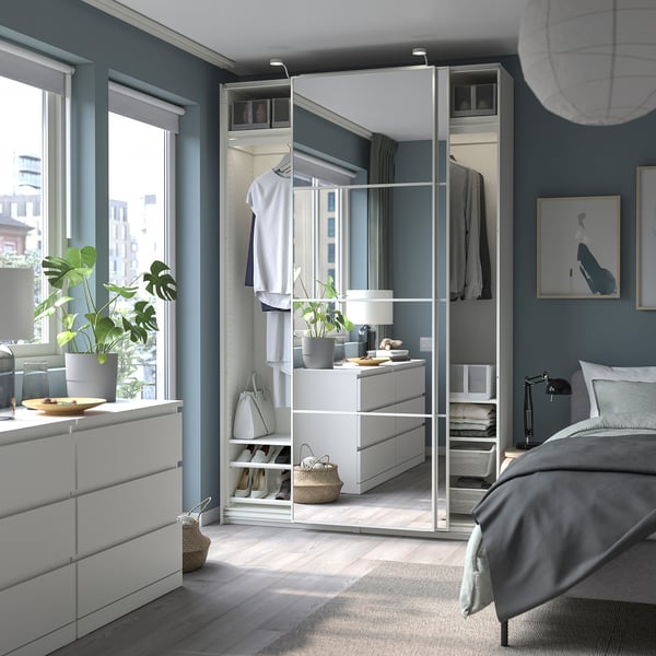 Modern bedroom with white PAX wardrobe, mirror, and sliding doors. Shelves, drawers, and hanging space. Blue-gray walls and large window.