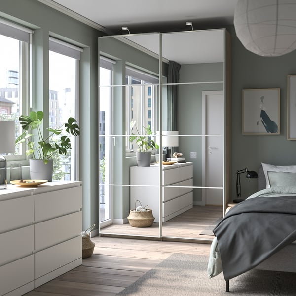 Modern bedroom with PAX wardrobes featuring mirrored sliding doors, grey bedding, white furniture, and indoor plants.