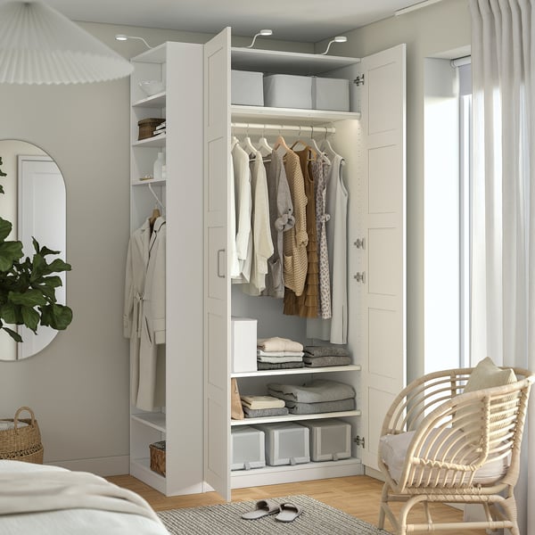 Bedroom with a white PAX wardrobe displaying clothes, shelves, and storage boxes, next to a mirror and a wicker chair.