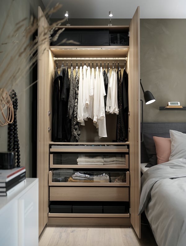Modern wooden wardrobe, open showing clothes and drawers. Dark wall, grey bed, wall lamp.