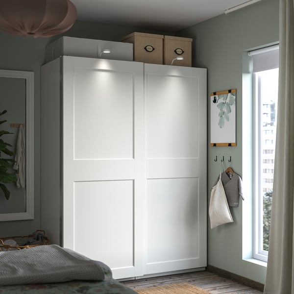 Modern bedroom with white PAX wardrobe, sliding doors, integrated lighting, and storage boxes.
