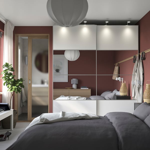 Modern bedroom with red walls, PAX white wardrobe, sliding doors, grey bed, hanging clothes, plants, and lamps.