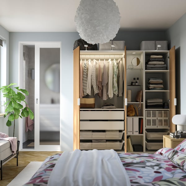 Bedroom with open wardrobe displaying hanging clothes, folded towels, shelves, and boxes. Next to a bed with a patterned blanket.