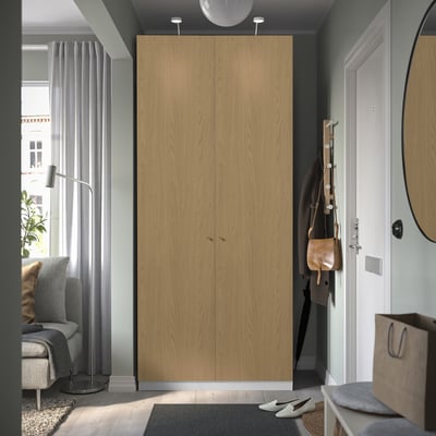Modern light brown PAX wardrobe near white door, taller than grey chair and lamp.