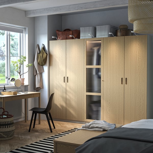 Modern bedroom with tall light-colored wooden PAX wardrobes, black chair, desk by window, striped rug.