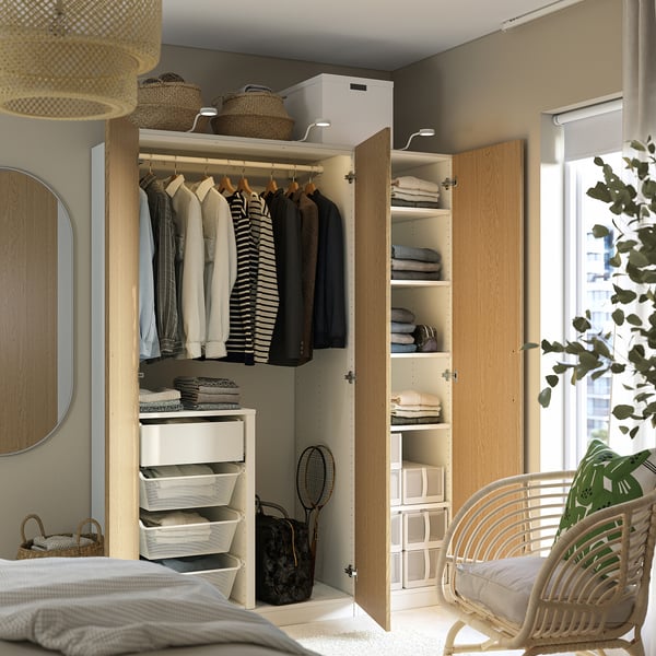 Modern bedroom with open PAX wardrobe showing clothes, shelves, and baskets; wicker chair and plant nearby.