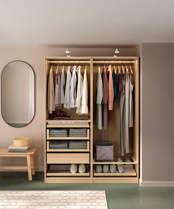 An open wooden PAX wardrobe displays neatly organised clothes and accessories. Light and airy, it glass organisers and drawners for storage.
