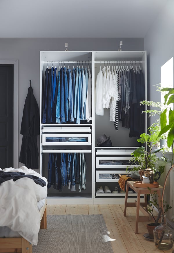 Modern bedroom with open PAX wardrobe, neatly arranged clothes, and a large plant.