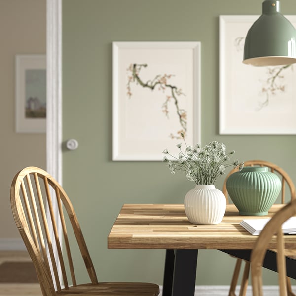 Cosy dining area with wooden table, PELARBJÖRK vase with flowers, under modern lamp, sage green wall with pictures.