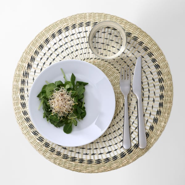 A round woven place mat with a grey and white pattern holds a white plate of salad and cutlery.