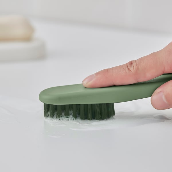 A hand holds a green oval brush with dark bristles, used for cleaning large surfaces and tight spaces.