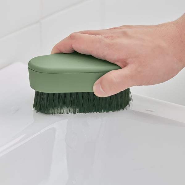 A hand holds a green PEPPRIG brush, scrubbing a white surface, showcasing its cleaning function.
