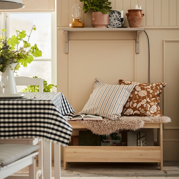 Cosy room with pine bench, floral pillows, plants, and chequered cloth.