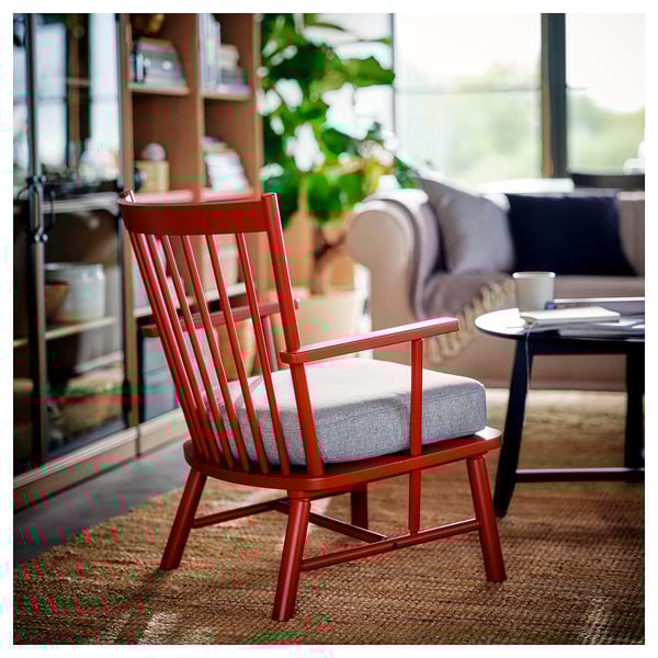 Red spindle armchair with cushion in cosy living room. Wooden frame, smaller than couch, with light cushion. Includes bookshelf, plants, and warm rug.