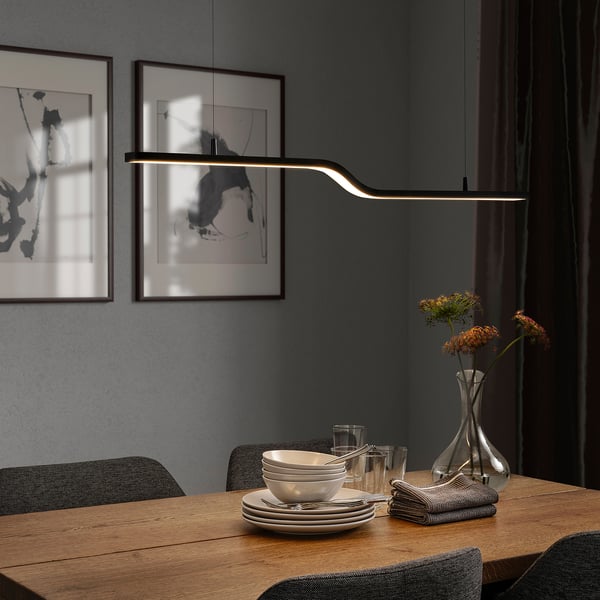 Modern dining room with PILSKOTT lamp, wooden table, & white dishes. Dark grey walls, dried flowers, & black/white art.