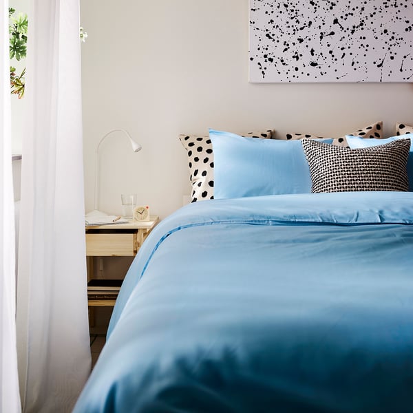 Bright blue PILTANDVINGE duvet set covers bed, matching pillows. Artwork above, light on bedside table.