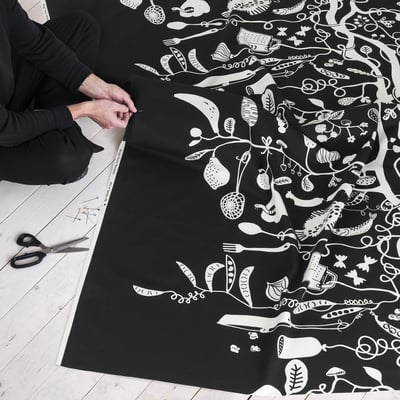 Person holds black fabric with white playful patterns, scissors and pins laid out, suggesting preparation for a sewing project.