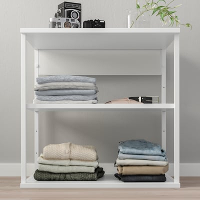 White PLATSA shelf unit with folded clothes, camera display, and plant.