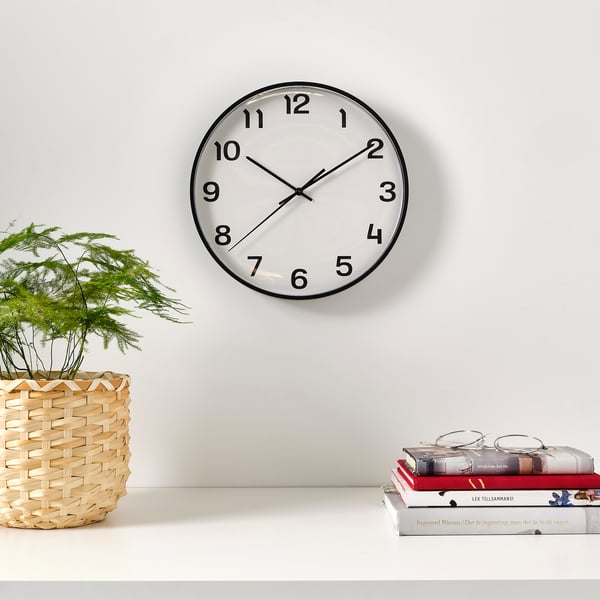A round wall clock, its face white with black numbers and hands, mounted above a shelf with books and a plant.