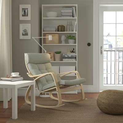 POÄNG rocking chair in calm setting. Light grey, high back, with side table and bookshelf. Neutral decor includes ottoman and curtains.