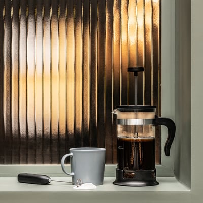 Black cafetière coffee maker on window sill with grey mug, frothing wand, and velvet bag near window with vertical blinds.