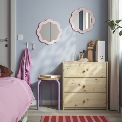 Pink-accented bedroom with blue walls, PRUNKHALLON mirrors, dresser, stool, rug, plant, and curtain.