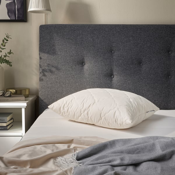 RÅDHUSVIN pillow in white on bed, cotton & wool, simple design. Bedside table with books & lamp beside it.