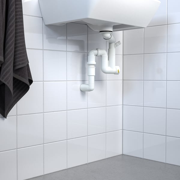 White sink with visible RÄNNILEN water trap and flexible piping against tiled wall.