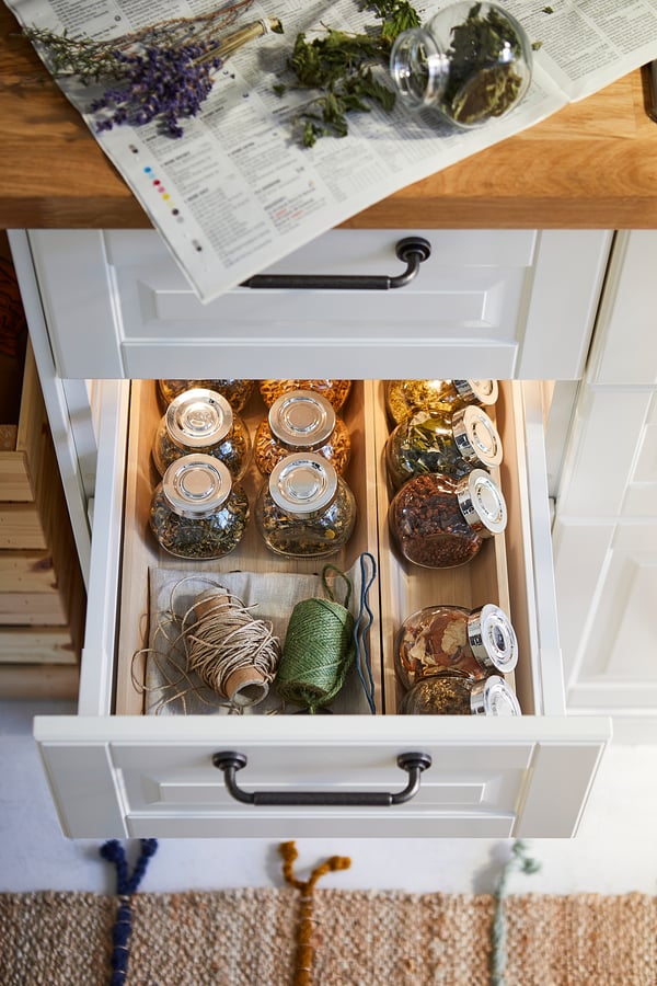 Open white drawer with glass jars storing herbs and strings, wooden countertop with herbs and a book above.