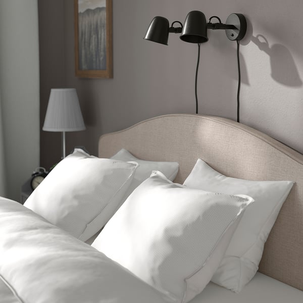 A bed with beige headboard, two white pillows, black wall light, and bedside lamp.