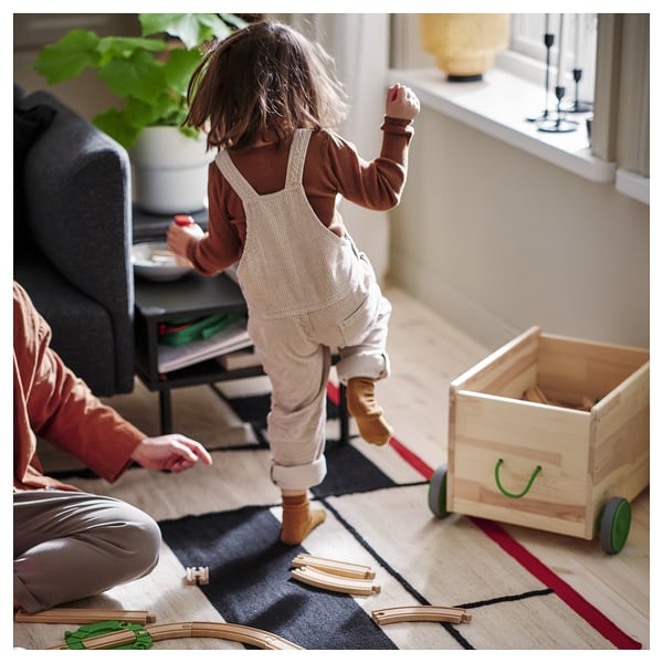 A kid in beige overalls plays with a toy train set on a beige and black rectangular rug with red edges, guided by an adult.