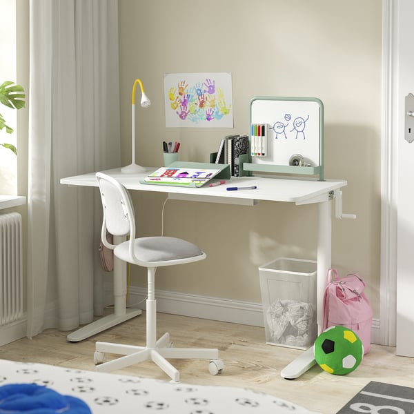 A white kids height-adjustable desk and chair with toys and school supplies, set against a light yellow wall.