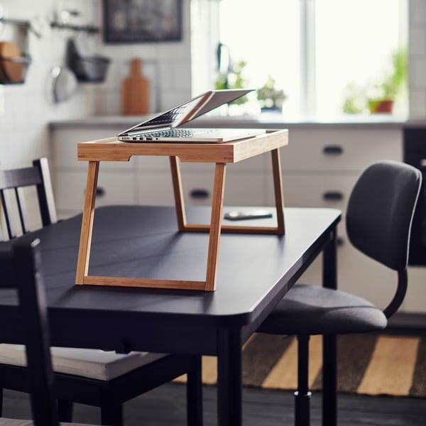 Wooden laptop table on a black table in a bright room, with a chair beside it.