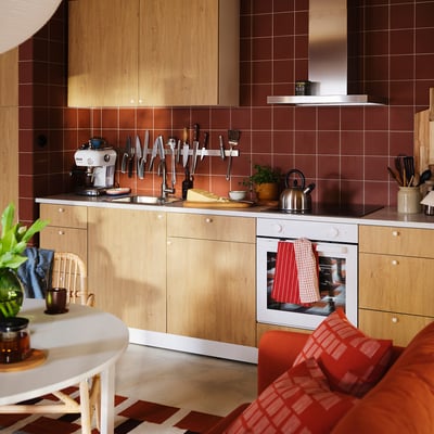 Kitchen with light wood cabinets, red tile backsplash, and cosy couch with chequered pillows. Red cotton RINNIG tea towel hangs neatly on oven handle.