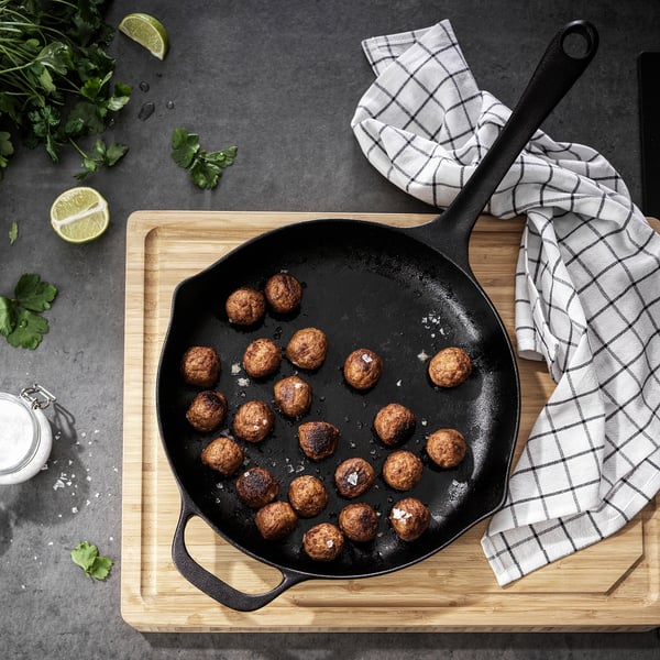 A cast iron skillet with meatballs, herbs, lime, and salt on a wooden board with a chequered towel.