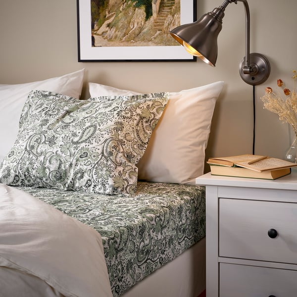 Bed with green paisley RODGERSIA sheets, cream pillows, nightstand with lamp, books, and vase. Fitted sheet stretches over large mattress.