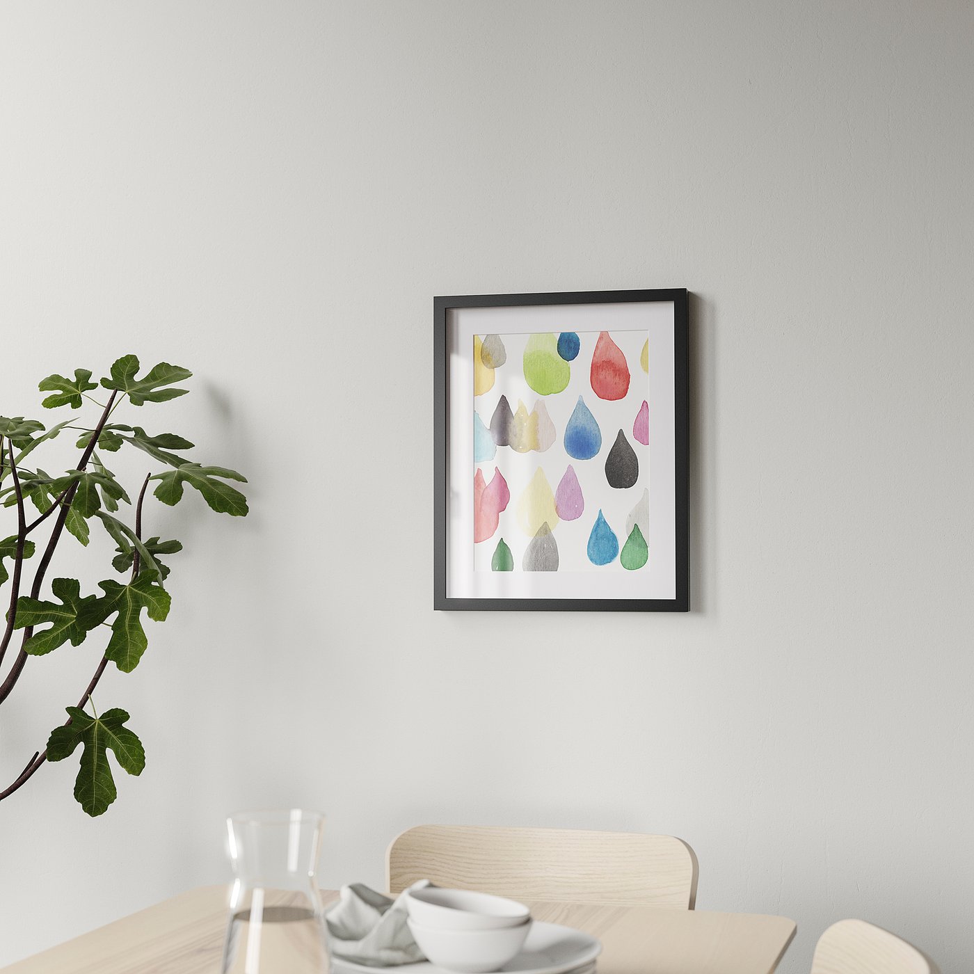 A modern black picture frame hangs on a wall above a dining table, showcasing colourful droplets and mounted with a neutral ph mount.