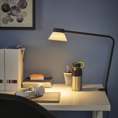 Desk-clamped SAMBORD lamp with triangular shade, illuminating workspace with books, bottle, and headphones.