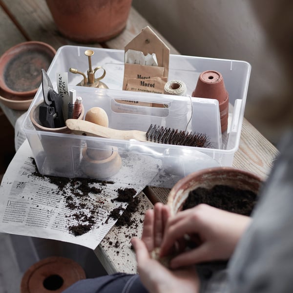 Clear plastic storage box with compartments, containing gardening tools and pots, placed on newspaper with soil.