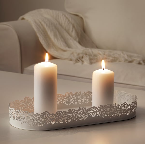 Two lit candles sit in an oval, white, laser-cut metal tray with leaf patterns, adding romantic shadows and ambiance to a light wooden table.