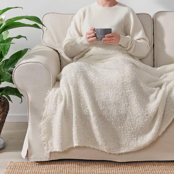 Person sitting on cream sofa, wrapped in a cosy, off-white SANDBRODD throw blanket, holding a mug.