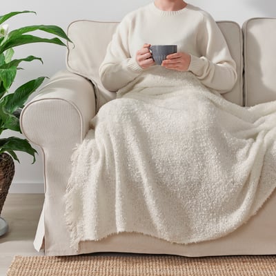 Person sitting on cream sofa, wrapped in a cosy, off-white SANDBRODD throw blanket, holding a mug.