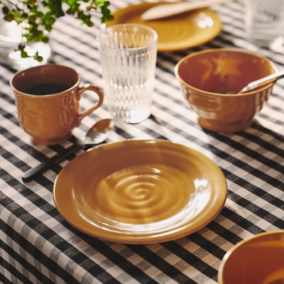 SANDSKÄDDA dinnerware in ochre yellow, including plates, cups, and a glass. Stoneware with rustic feel, stacked on chequered tablecloth.