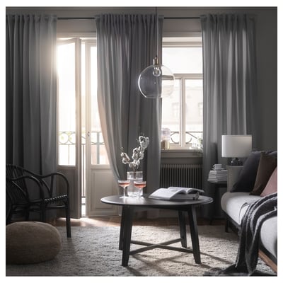 Modern living room with dark grey curtains, wicker chair, wooden table, wine glasses, and rug. Large window with soft light.
