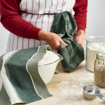 Person in red and striped apron handling green towel beside a mixing bowl with flour and measuring cup with grains.
