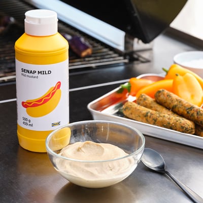Image a bottle of yellow mustard labelled SENAP MILD next to a bowl of sauce and a tray of sausages on a grill.