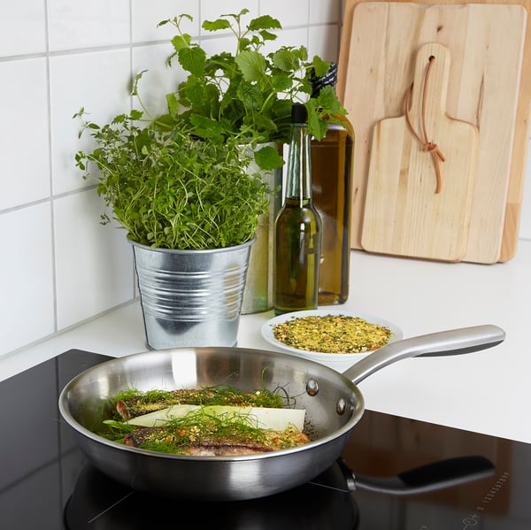 Stovetop with metal pan, herbs, butter, and fish. Wooden board and spices nearby. Ideal for cooking.