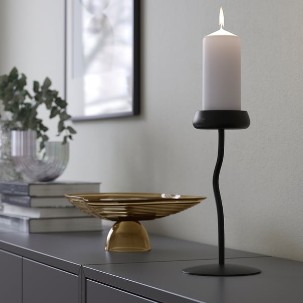 A modern black candlestick holds a white lit candle on a dark surface, offering mood lighting.